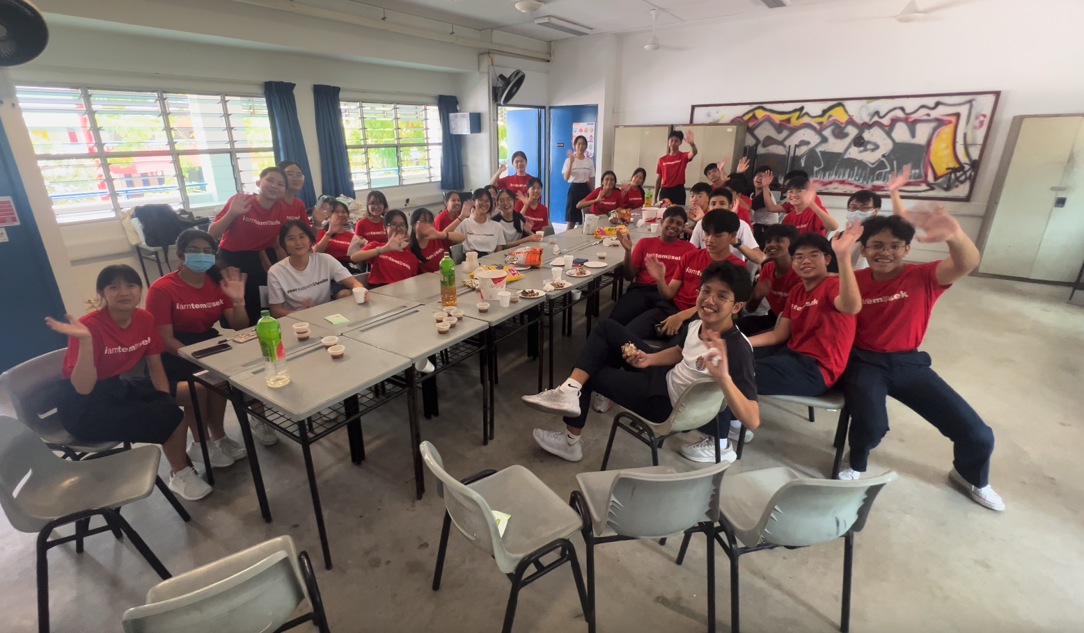 A large group of students in red Temasek shirts wave and smile around tables with snacks in a school room.
